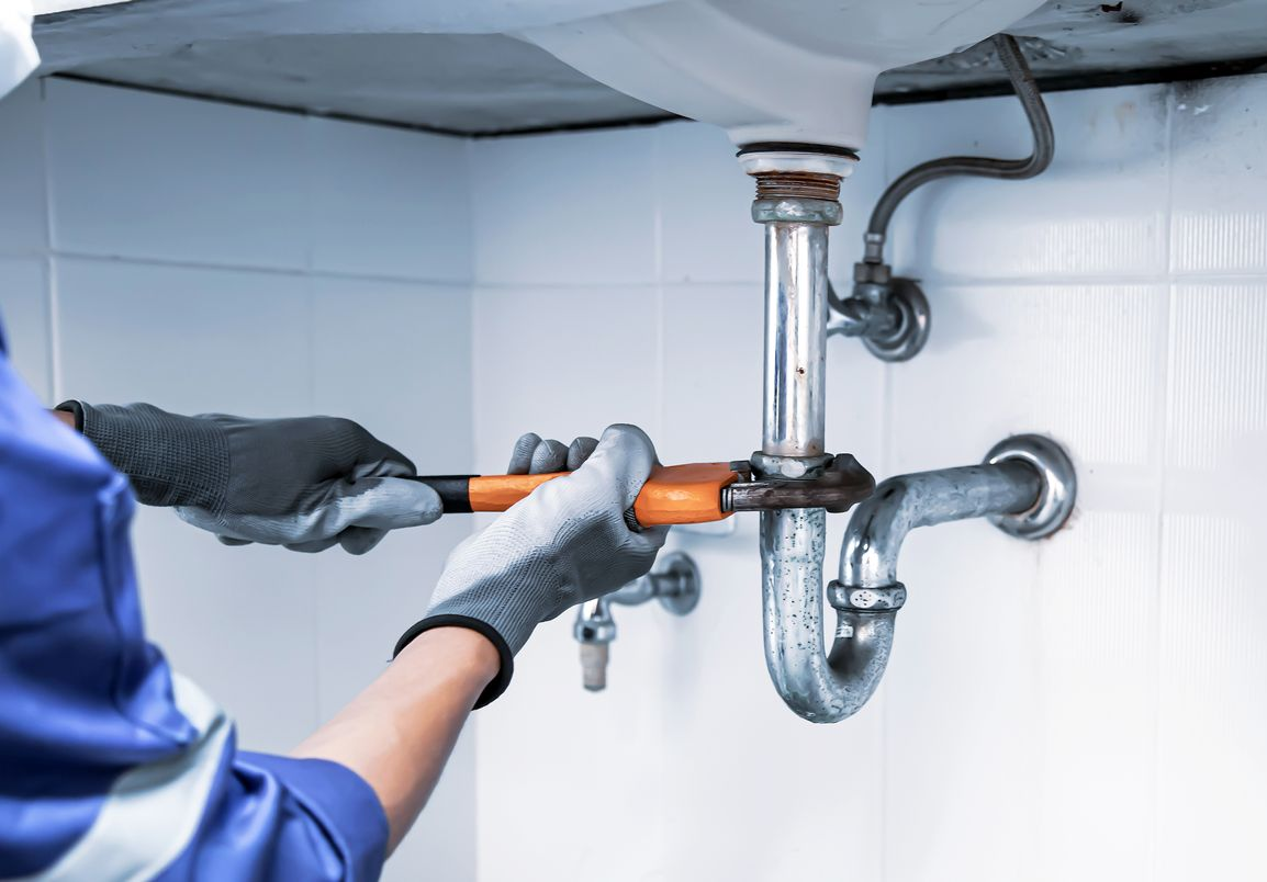 Un plombier dans une salle de bains, utilisant une clé pour travailler sur des tuyaux sous un lavabo.