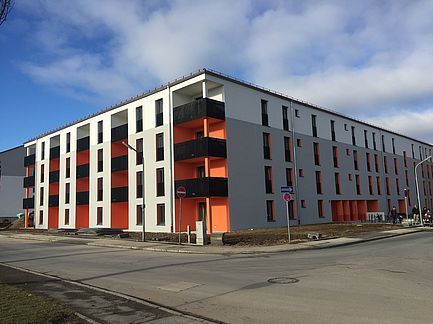 Modernes Wohnhaus mit orangefarbenen Balkonen an einer Straßenecke unter blauem Himmel