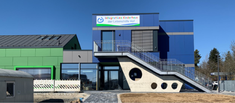 Modernes blaues Gebäude mit Außentreppe und Schild, neben einem grünen Gebäude unter klarem Himmel
