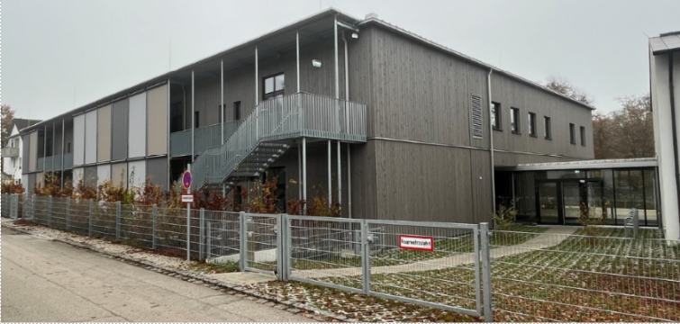 Modernes, graues zweistöckiges Gebäude mit Treppe, umzäuntem Innenhof und kahlen Bäumen an einem bewölkten Tag