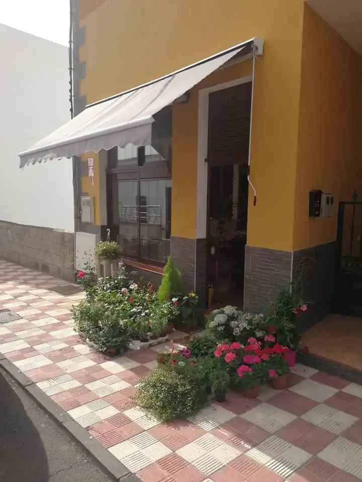 Un edificio amarillo con toldo, puerta y flores en macetas, sobre una acera a cuadros rojos y blancos.