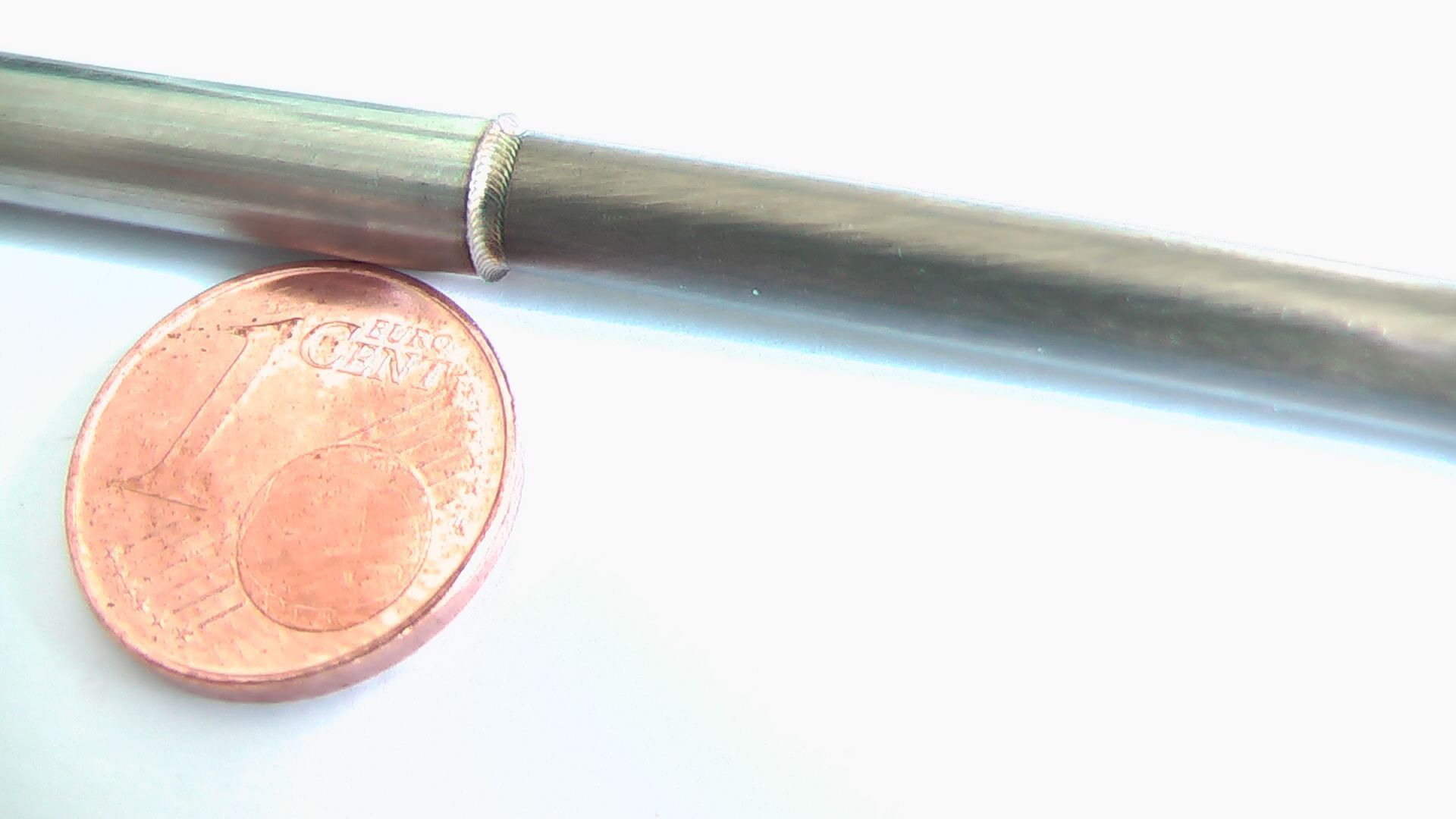 Eine Kupfermünze liegt neben einem Metallrohr auf einer weißen Oberfläche um das Laserschweißen zu demonstrieren.