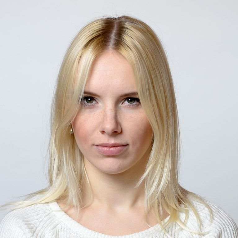 Mujer rubia de piel clara, ojos claros y expresión neutra, vestida con un suéter blanco, sobre un fondo gris claro.