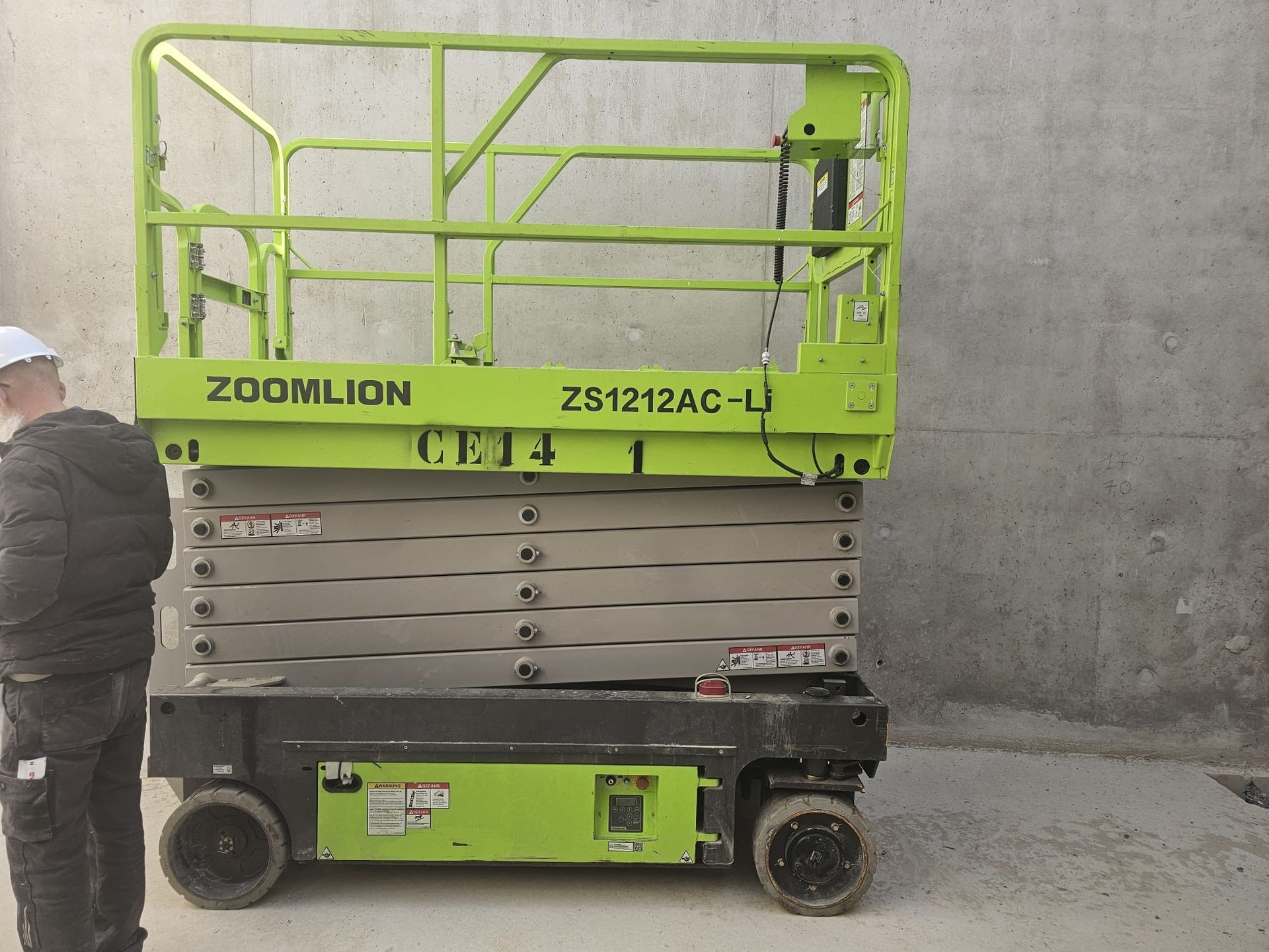 Une nacelle élévatrice à ciseaux Zoomlion ZS1212AC-Li vert citron est garée contre un mur en béton gris près d'une personne en tenue de travail.