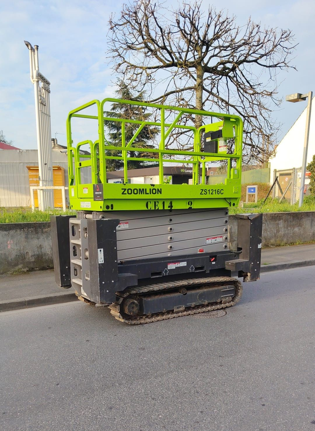 Une nacelle élévatrice à ciseaux vert vif, montée sur chenilles, est stationnée sur une route asphaltée grise sous un ciel d'un bleu éclatant.