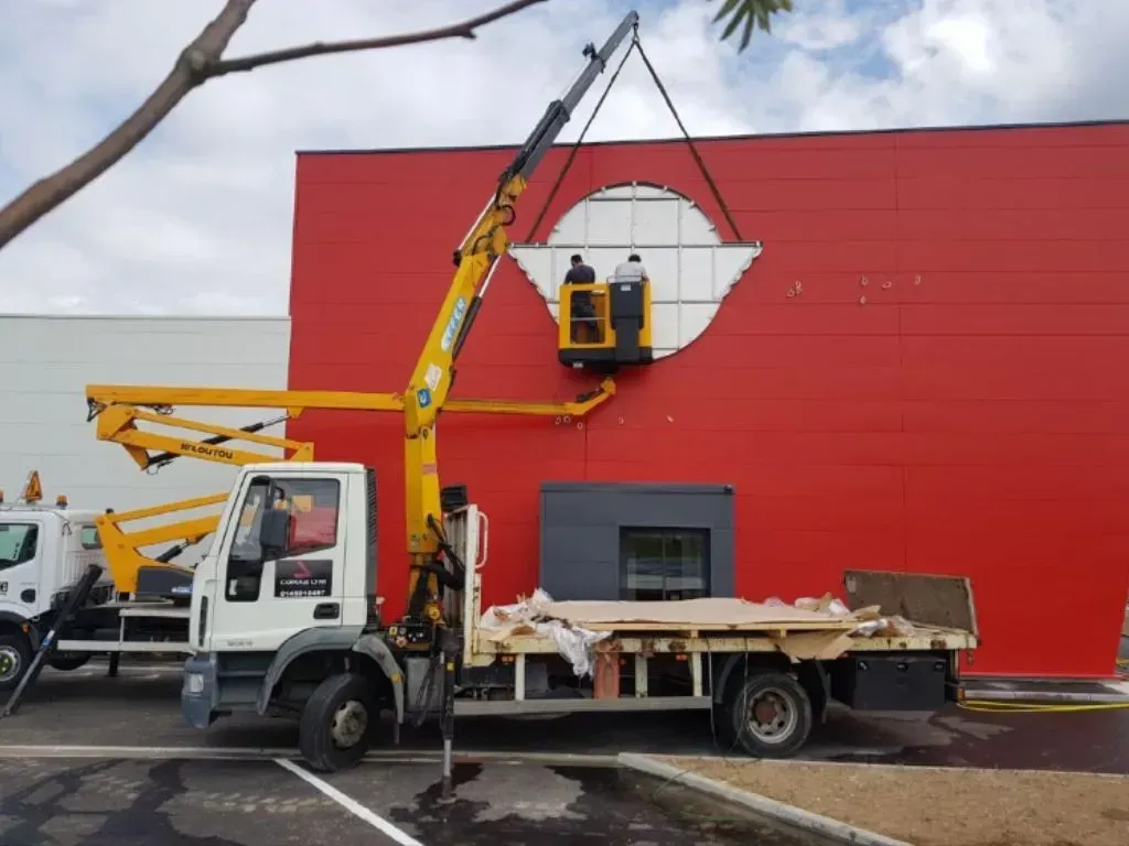 Un camion-grue soulève deux ouvriers dans une nacelle pour installer un panneau circulaire blanc sur un mur extérieur rouge vif.