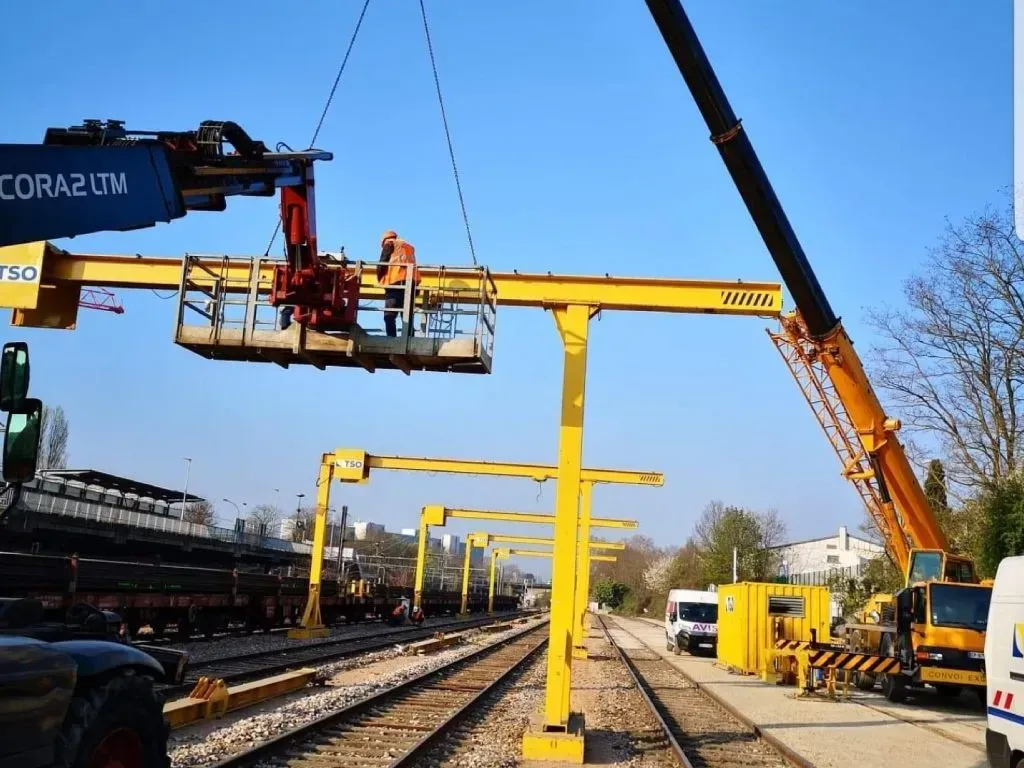 Une grue mobile soulève un ouvrier sur une plateforme pour entretenir une structure de portique ferroviaire le long des voies.