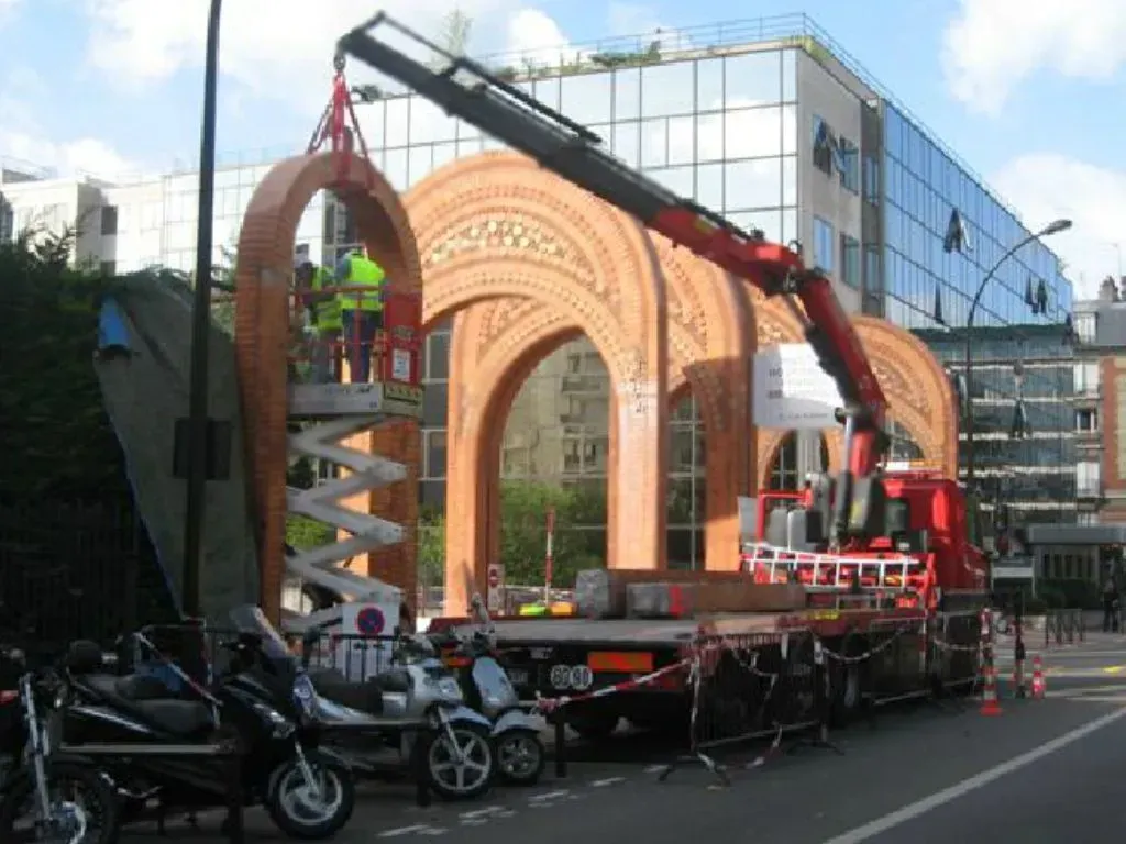 Un camion-grue rouge soulève un segment d'arche en briques décoratives dans une rue de la ville, tandis que des ouvriers se trouvent à proximité sur une nacelle élévatrice.