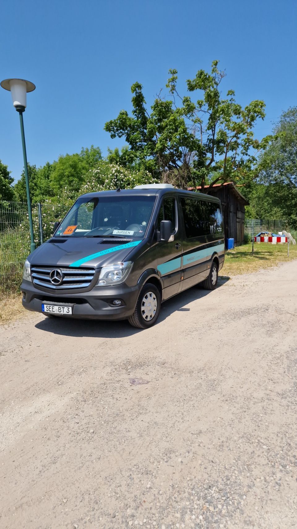 An einem sonnigen Tag parkte ein grauer Mercedes Sprinter-Van auf einer Schotterstraße neben einem kleinen Gebäude.