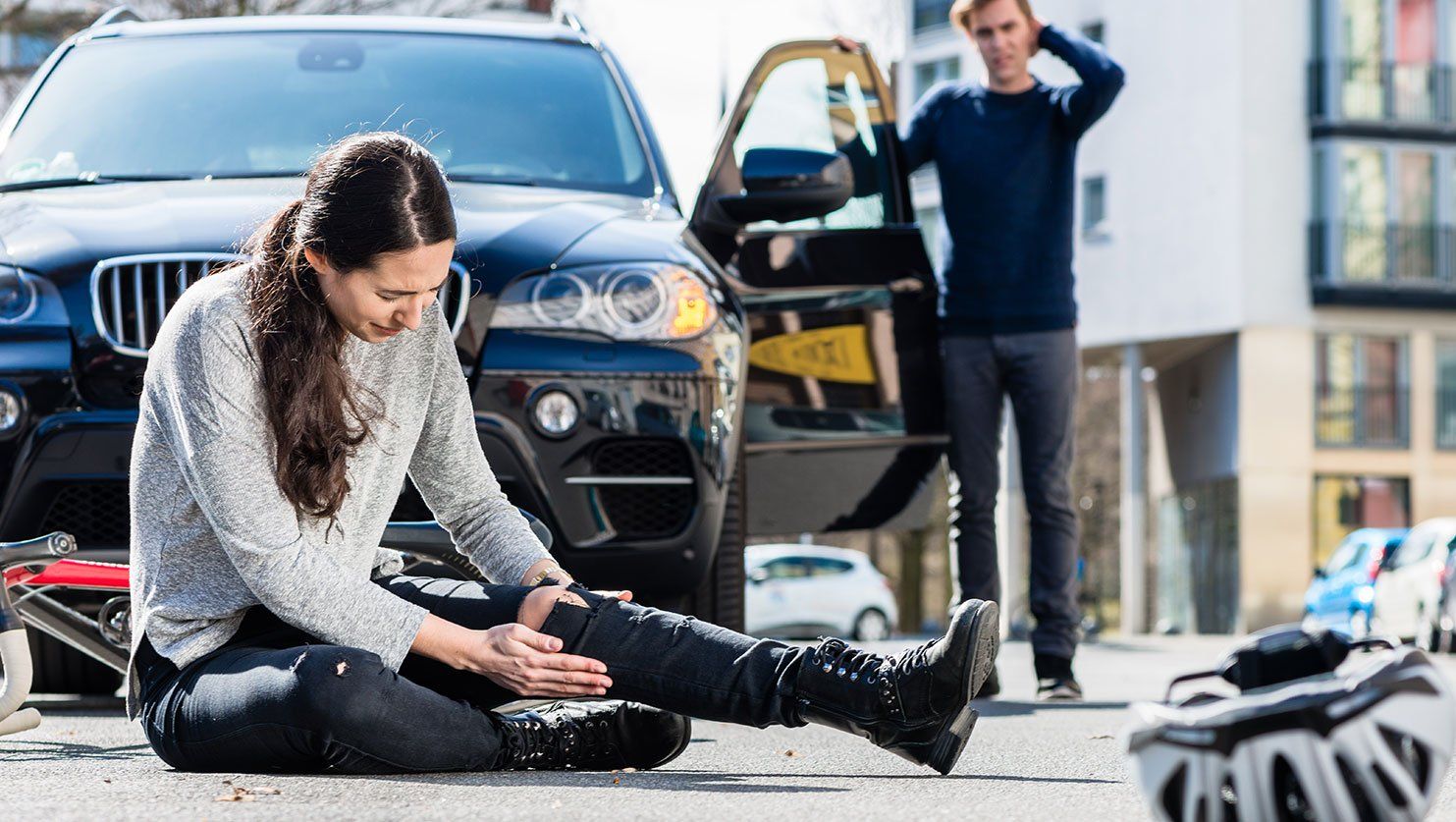 Une femme se tient la jambe au sol à côté de son vélo, un homme sort de sa voiture à l'arrêt