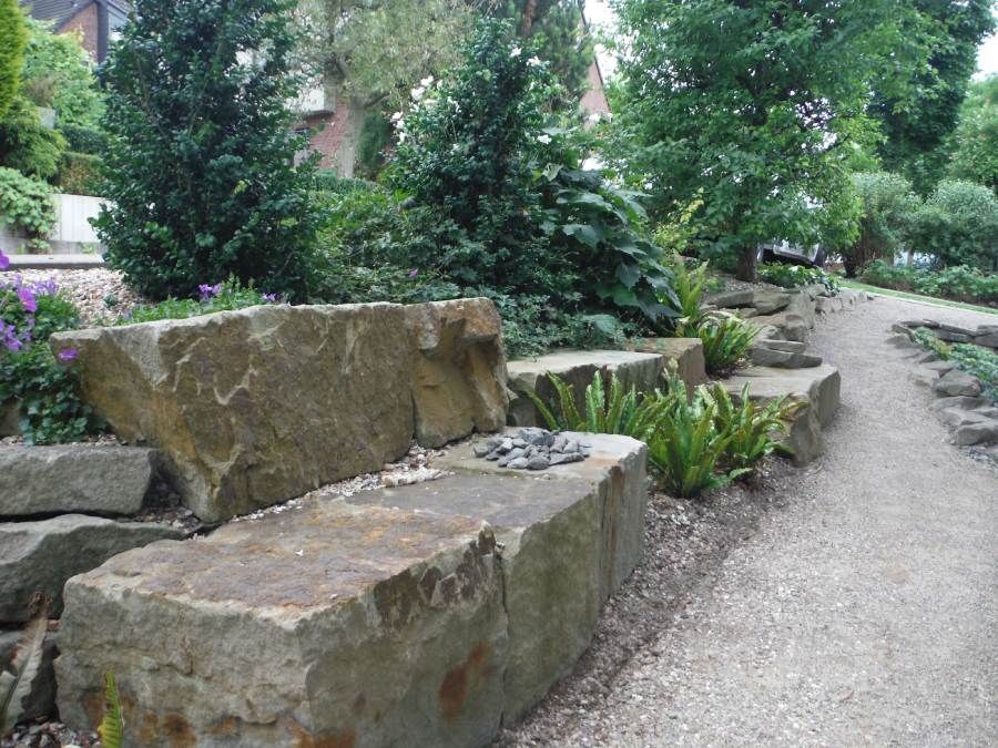 Steinmauer mit Pflanzen, Kiesweg und Bäumen in einer Gartenanlage.