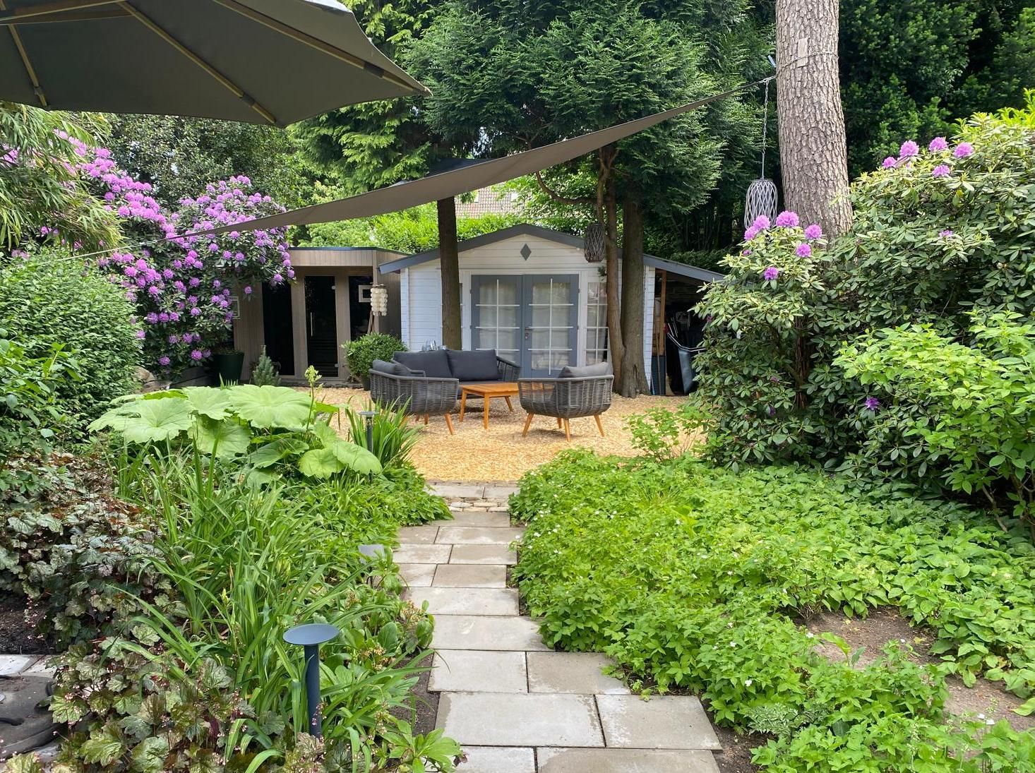 Ein Steinweg führt zu einem Garten mit einem weißen Schuppen, einer Sitzecke, üppigem Grün und teilweisem Schatten.