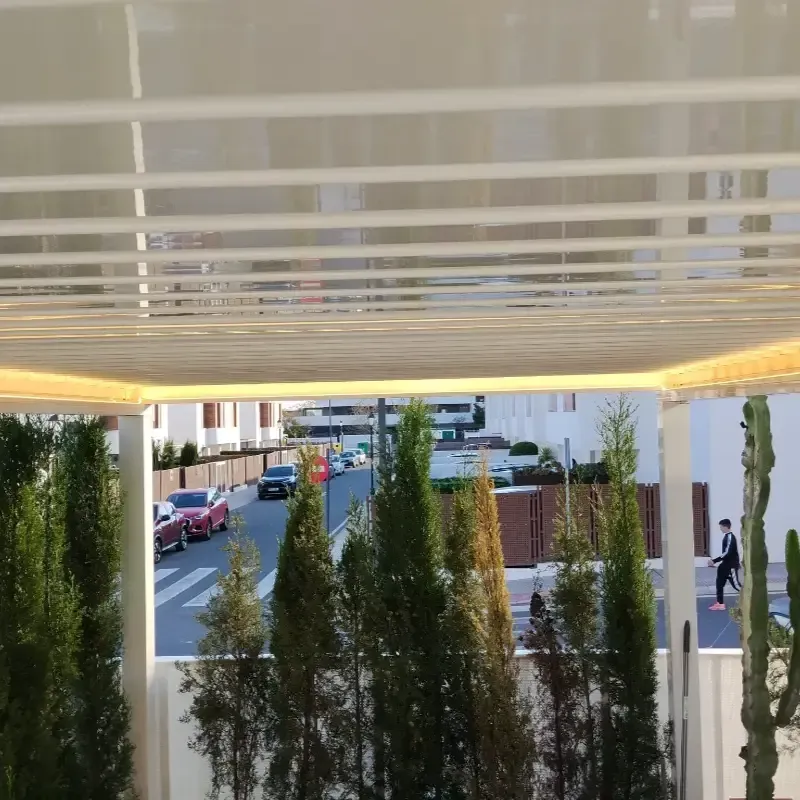 Vista desde debajo de un toldo blanco con altas plantas verdes que enmarcan una calle y peatones en la distancia.