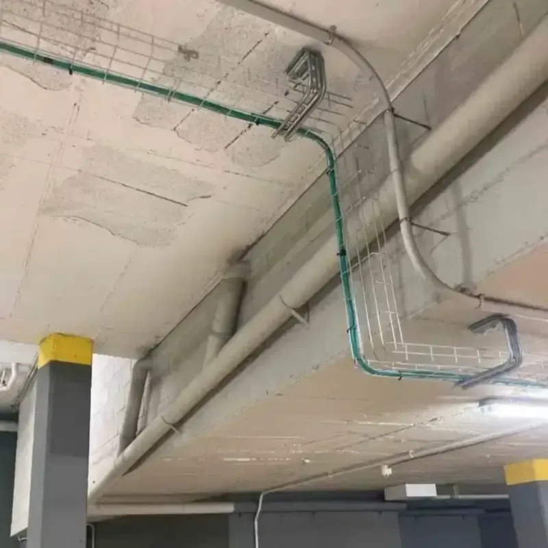 Techo industrial con conductos grises, canalizaciones verdes e iluminación fluorescente en un estacionamiento.