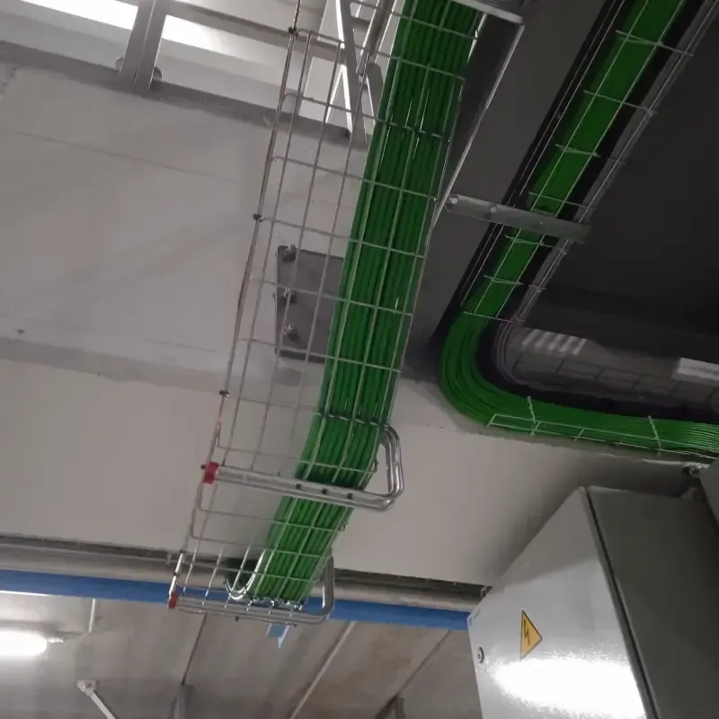 Bandejas portacables y tuberías de color verde recorren un techo industrial junto a una unidad eléctrica gris.