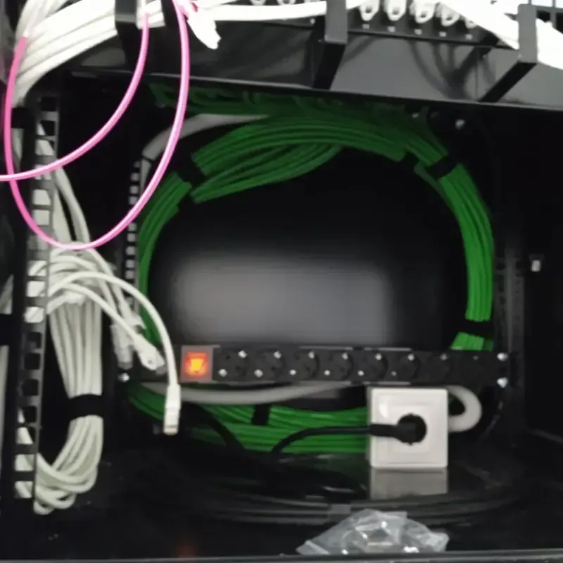 Rack de red con dispositivo negro, cable en espiral verde y regleta de toma de corriente blanca.