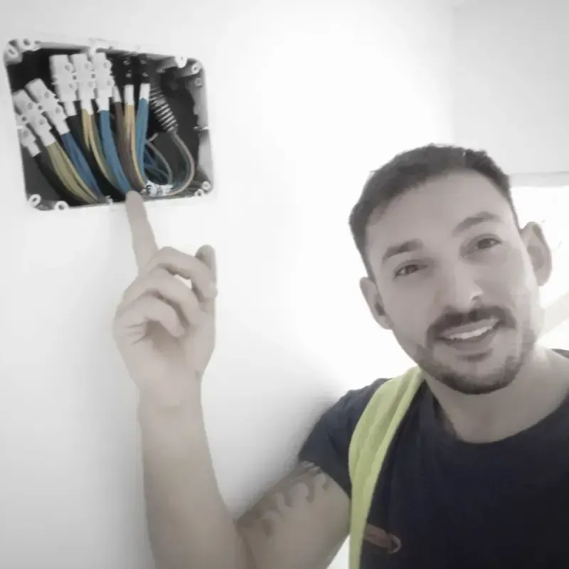 Un hombre sonríe y señala una caja eléctrica abierta con cables de colores en la pared.