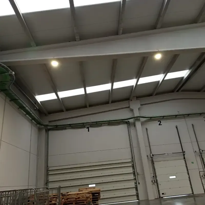 Interior de una nave industrial con techo alto de metal, luces cenitales y grandes puertas de carga.