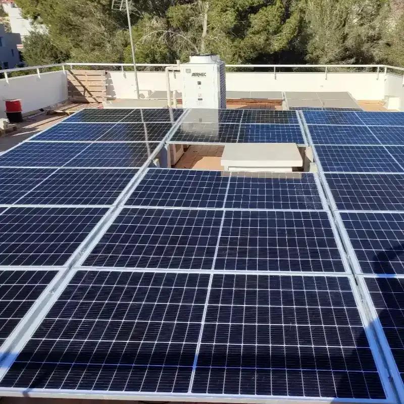 Paneles solares que cubren un tejado plano con una caja de servicios blanca y árboles al fondo.