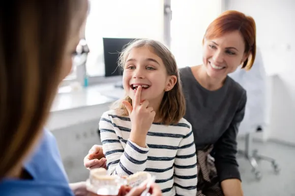 Niña en una cita con el dentista con su madre, señalando un diente mientras el dentista sostiene un modelo, sonríe.