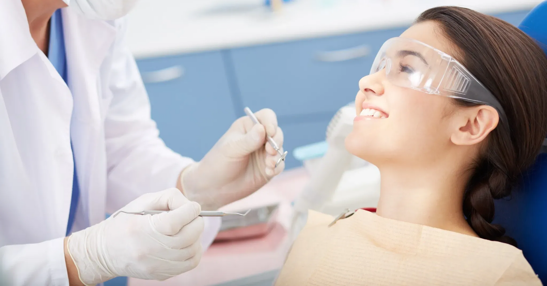 Dentista examinando los dientes de un paciente. El paciente usa gafas de seguridad y sonríe. En el entorno clínico.