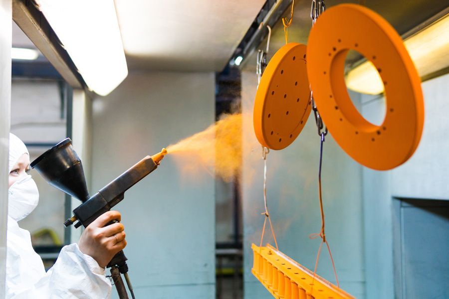 Une personne applique une peinture en poudre orange au pistolet sur des pièces métalliques dans un atelier, portant une combinaison et un masque de protection.