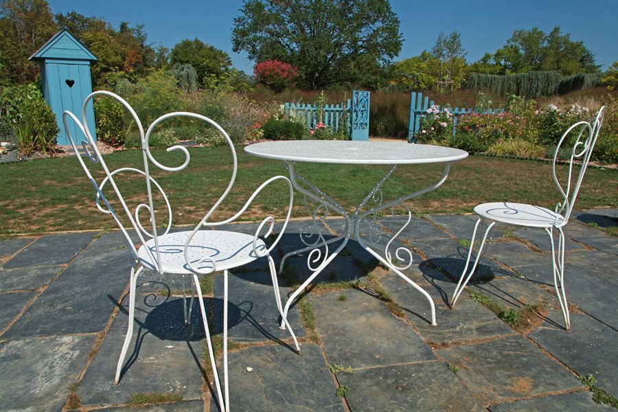 Table et chaises en fer forgé blanc sur une terrasse en pierre dans un jardin. Abri de jardin et portail bleus en arrière-plan.