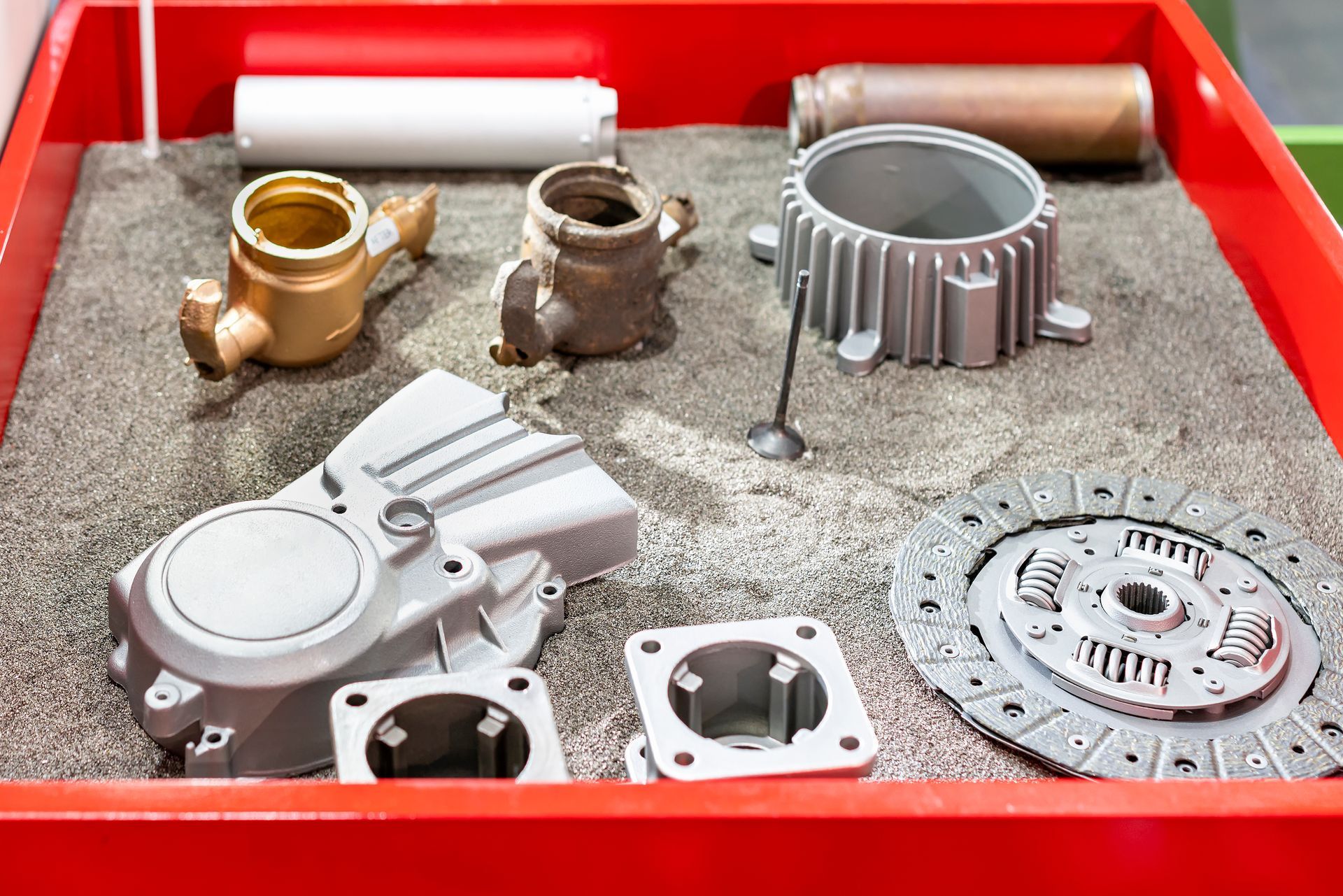 Pièces métalliques dans un bac rouge : un carter de moteur, un embrayage et d’autres composants dans le sable.