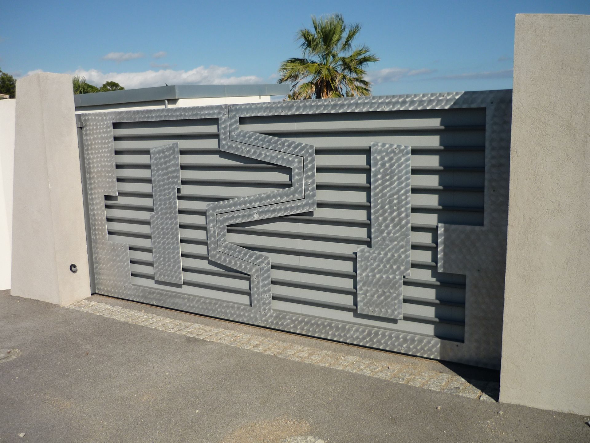 Portail en métal gris à motif géométrique, enchâssé dans des piliers en béton sur fond de ciel ensoleillé.