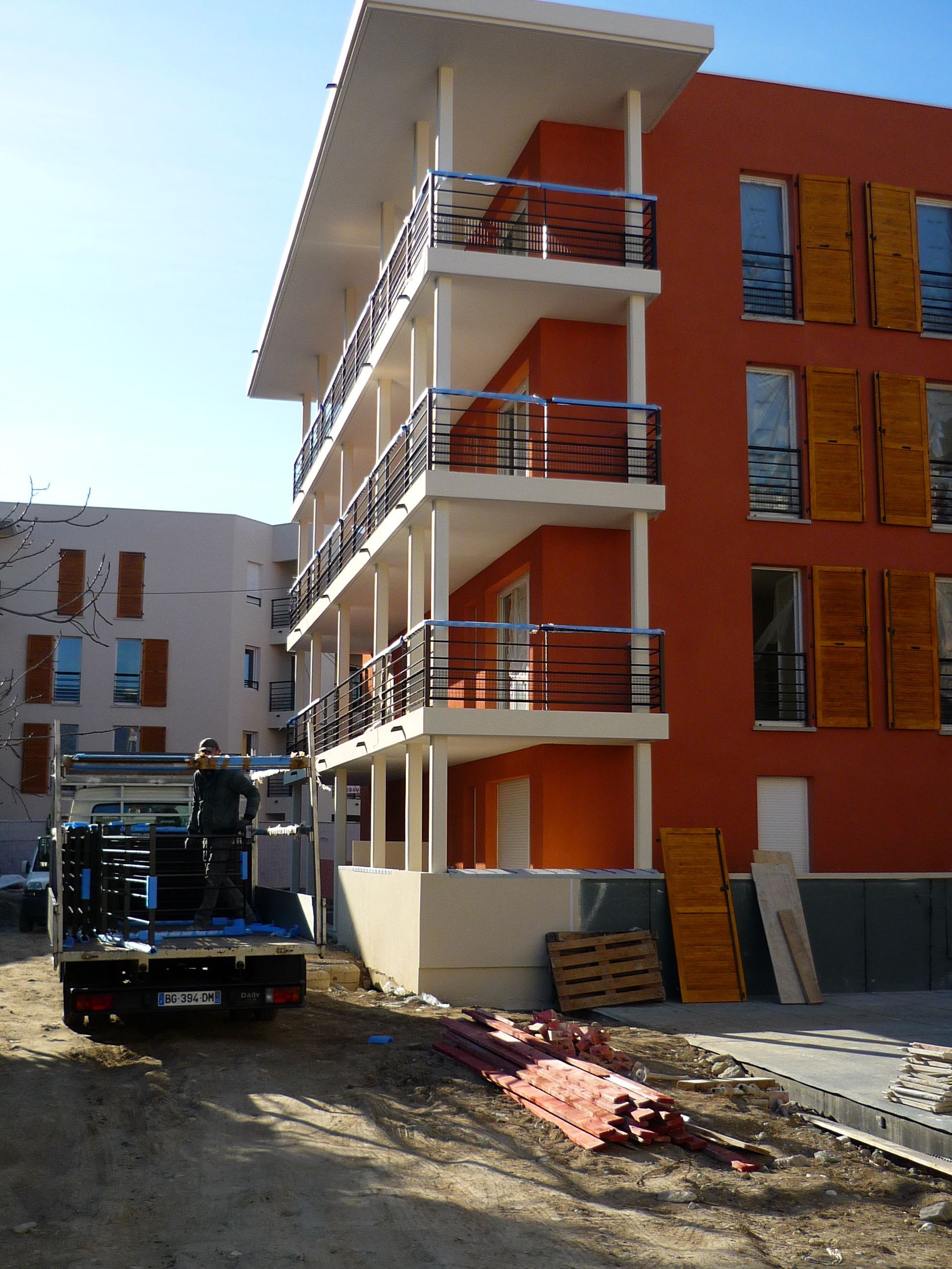 Un camion de livraison devant un immeuble d'appartements orange moderne avec des balcons, des matériaux de construction au sol.
