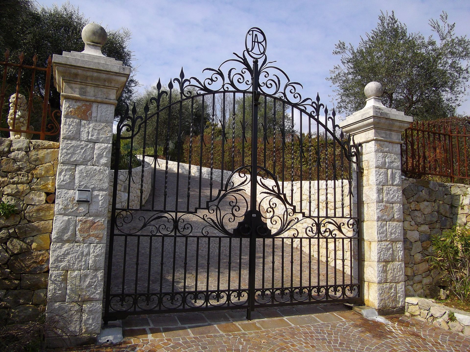 Un portail en fer forgé noir, entre des piliers de pierre, mène à une allée carrossable, se détachant sur un ciel lumineux.
