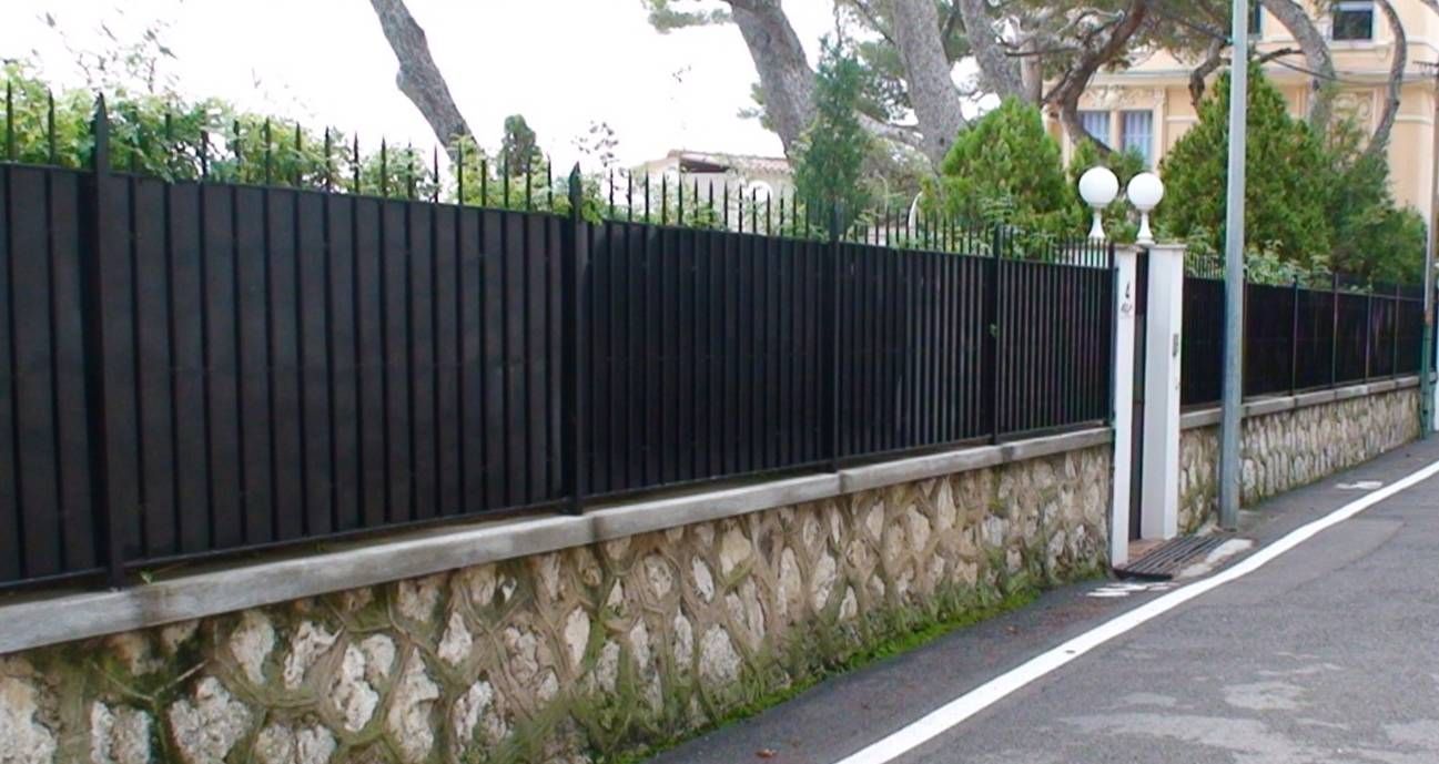 Clôture en métal noir avec socle en pierre, le long d'une route à bande blanche.