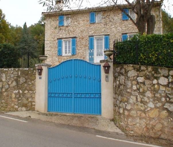 Maison en pierre avec volets et portail bleus ; mur en pierre, en bord de route.