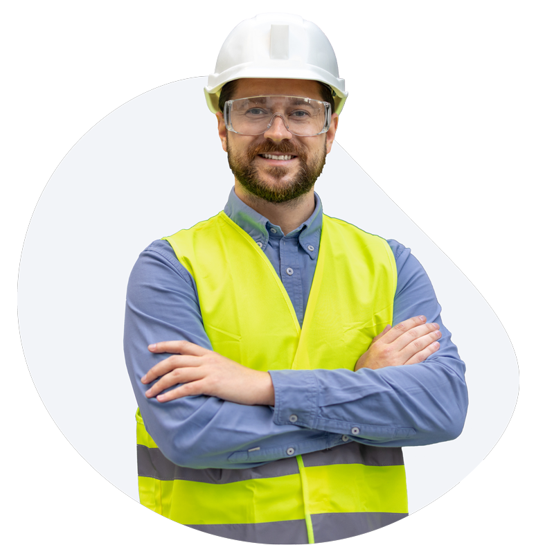Un homme portant un casque de chantier, des lunettes de sécurité et un gilet jaune, les bras croisés, sourit.