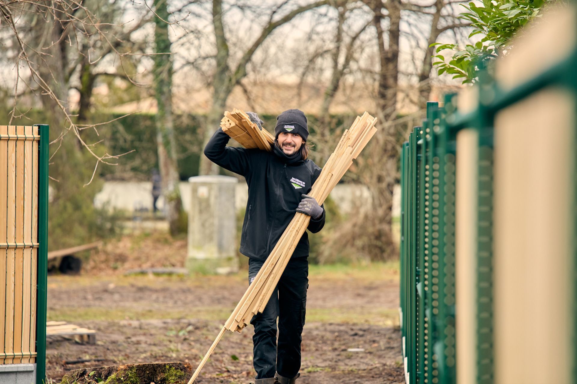 Un homme transporte des planches pour une clôture à l'extérieur ; il porte un chapeau, des gants et une veste noire, et il sourit.