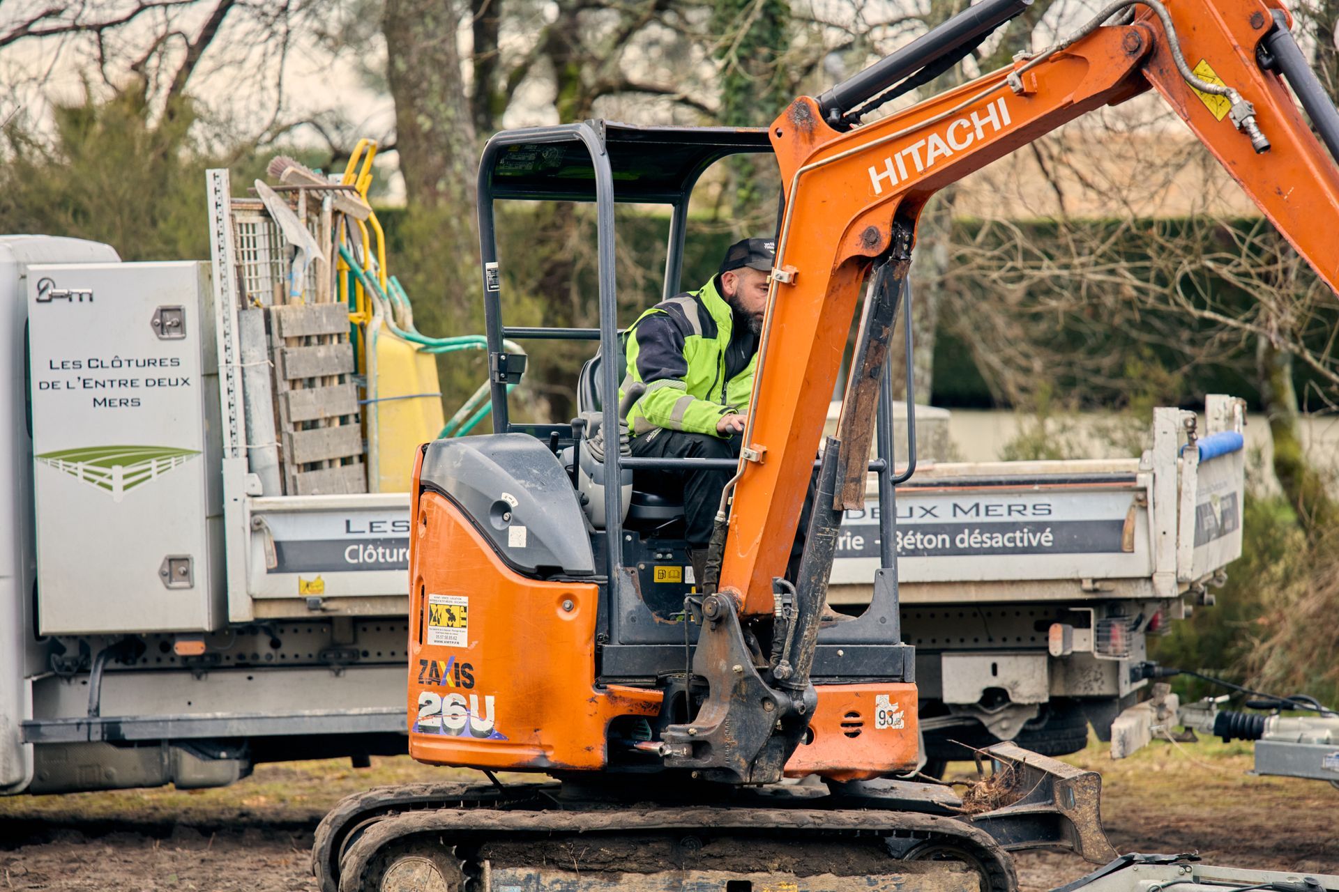 Une personne manœuvrant une pelle mécanique Hitachi orange près d'un camion blanc.