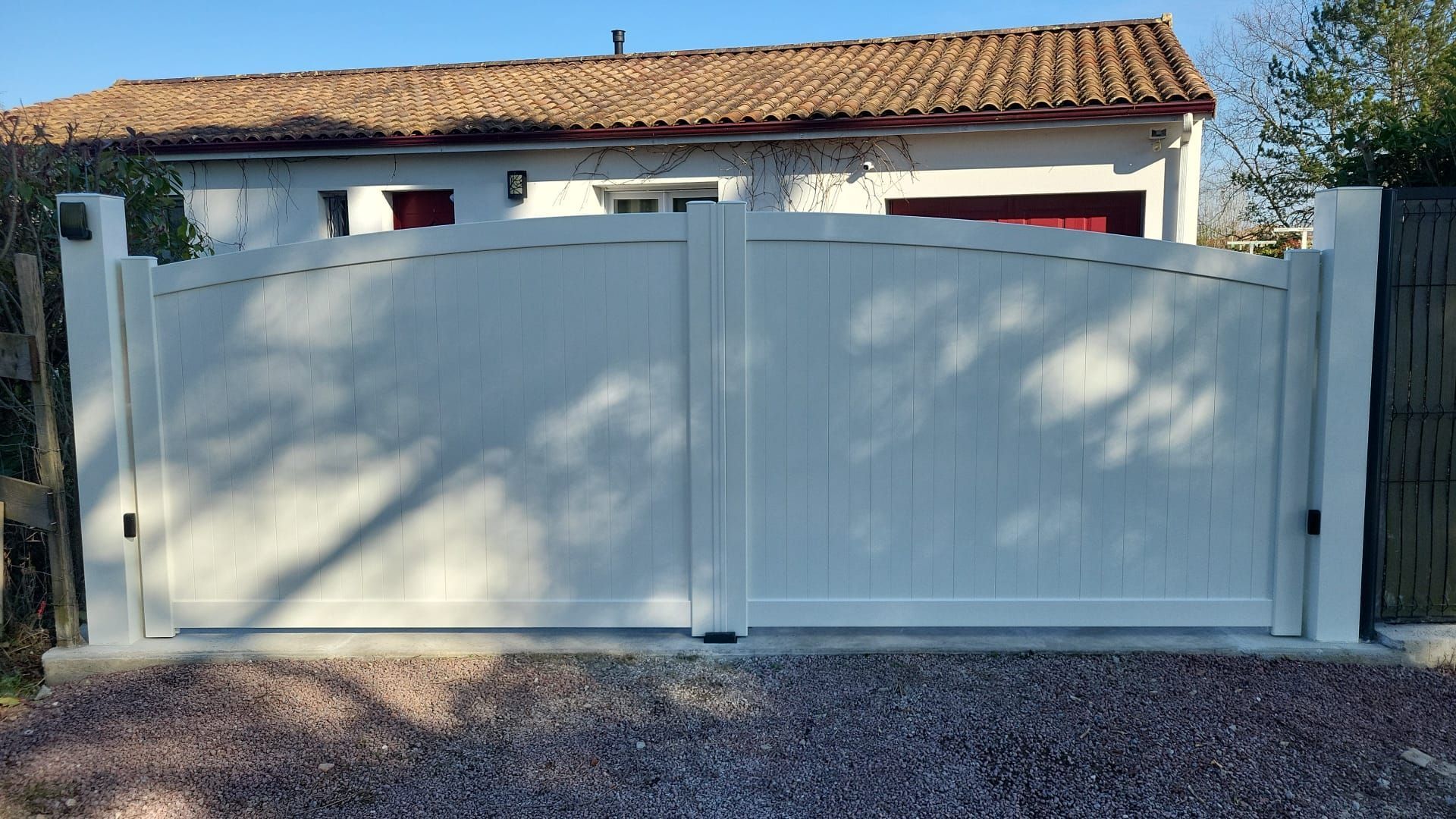 Portail d'allée blanc en arc de cercle, fermé. Allée de gravier au premier plan, maison au toit de tuiles à l'arrière-plan.