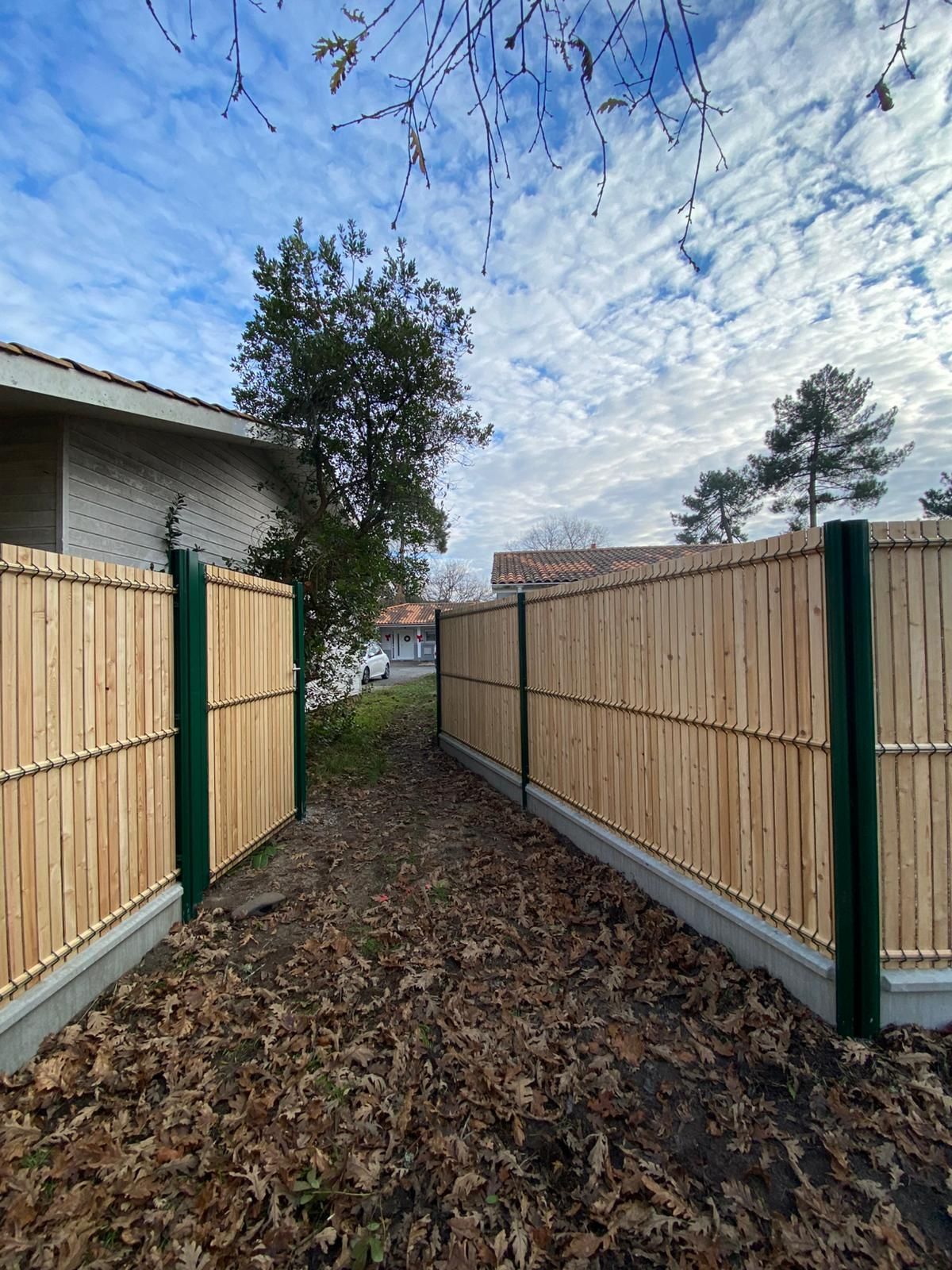 Clôture en bambou aux poteaux verts, menant à un sentier sous un ciel nuageux.