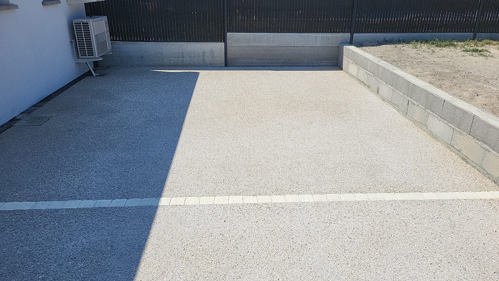 Espace extérieur recouvert de gravier, bordé de béton et avec une clôture noire en arrière-plan.