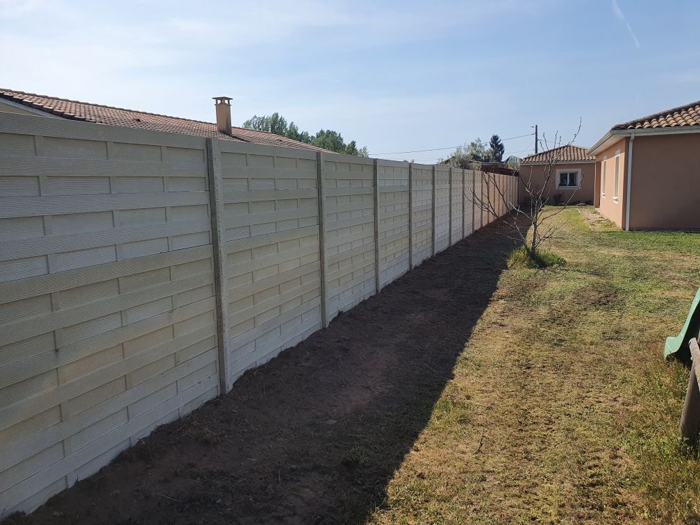 Clôture en béton gris dans une cour arrière, avec un chemin de terre le longeant et de l'herbe de chaque côté.