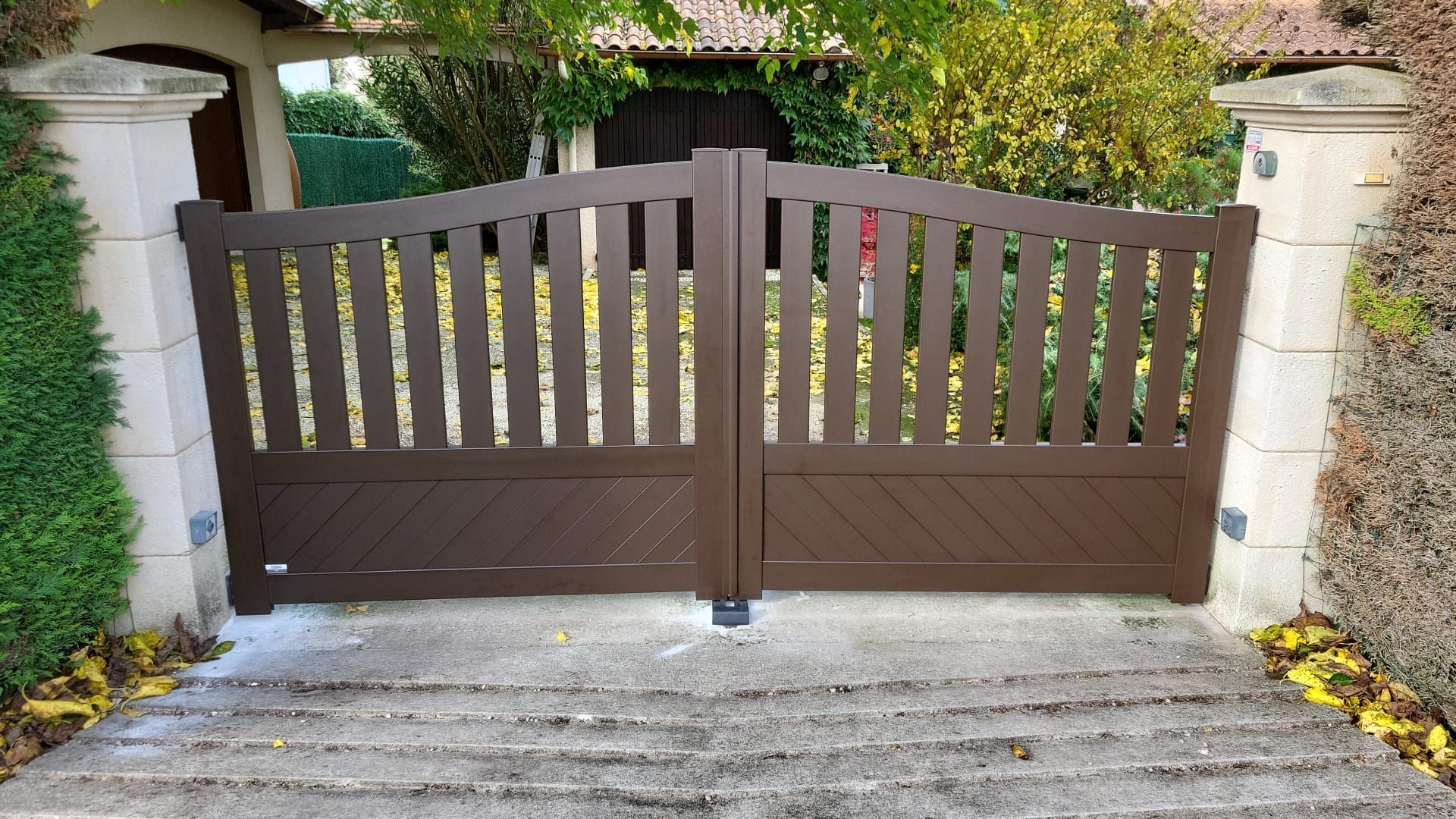 Portail double en bois brun à lattes verticales, placé entre des colonnes en pierre sur une allée.