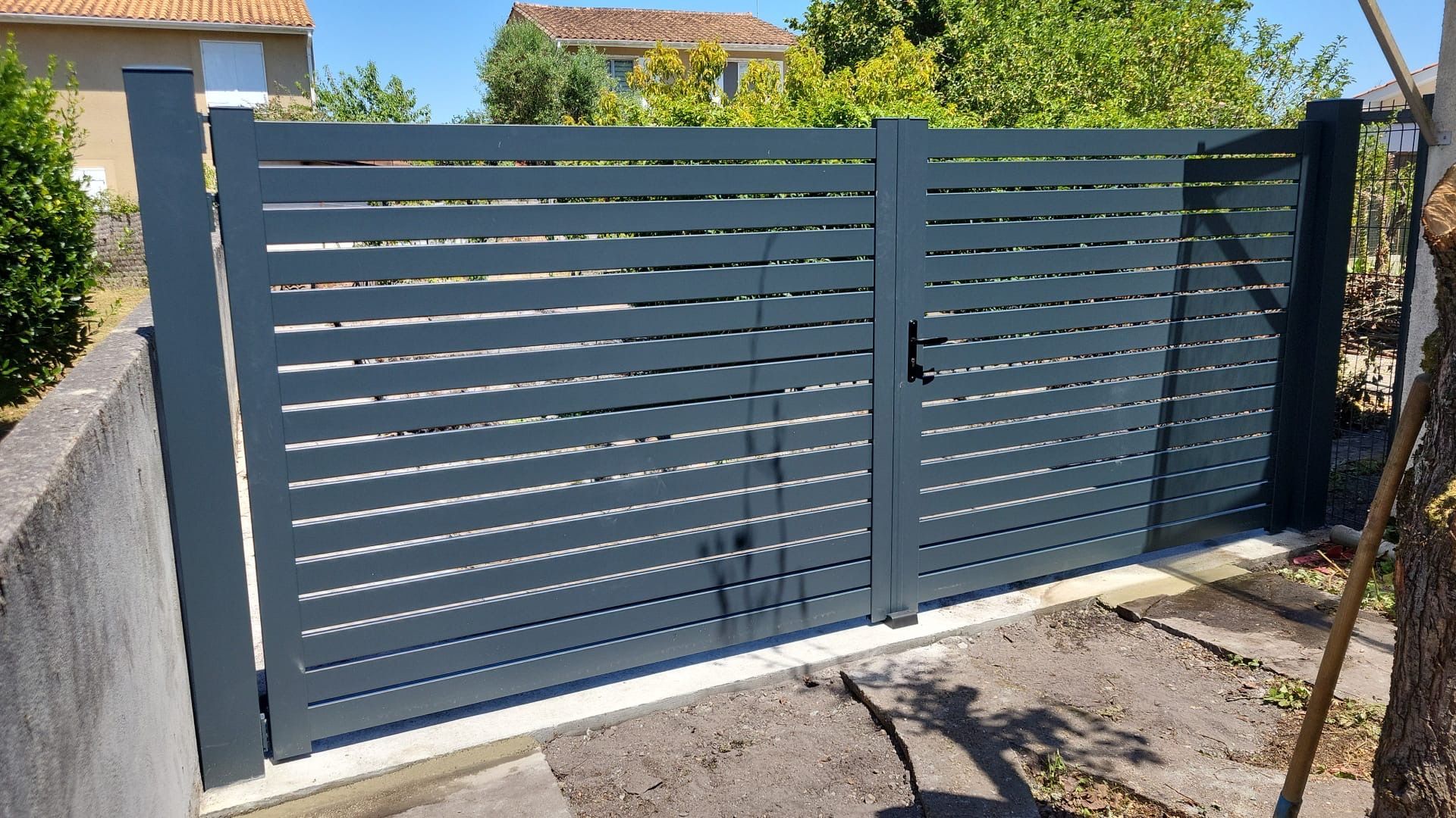 Portail métallique gris à lattes horizontales, devant une maison.