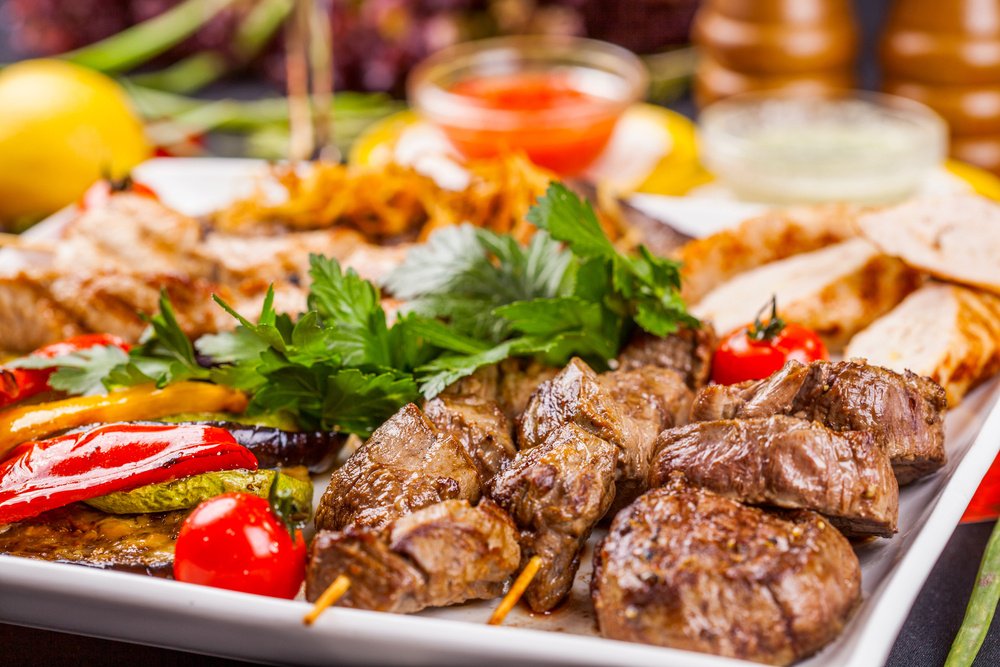 Brochettes de viande grillée aux poivrons et aux herbes sur un plat blanc, avec divers plats en arrière-plan.