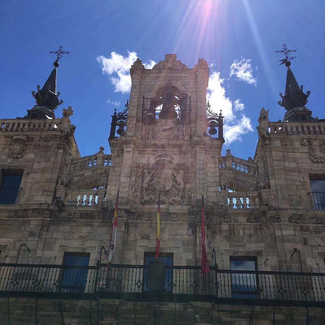 Fachada de catedral de piedra ornamentada con dos agujas bajo un cielo azul brillante y un destello de sol.