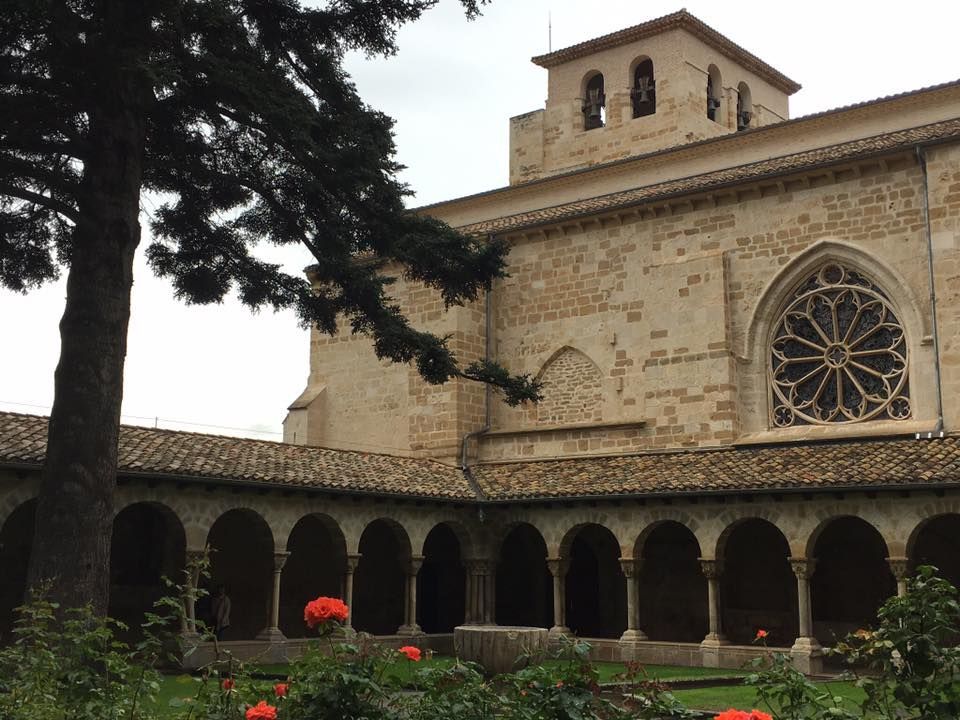 Claustro de piedra con columnata arqueada, rosetón y un árbol en el patio.