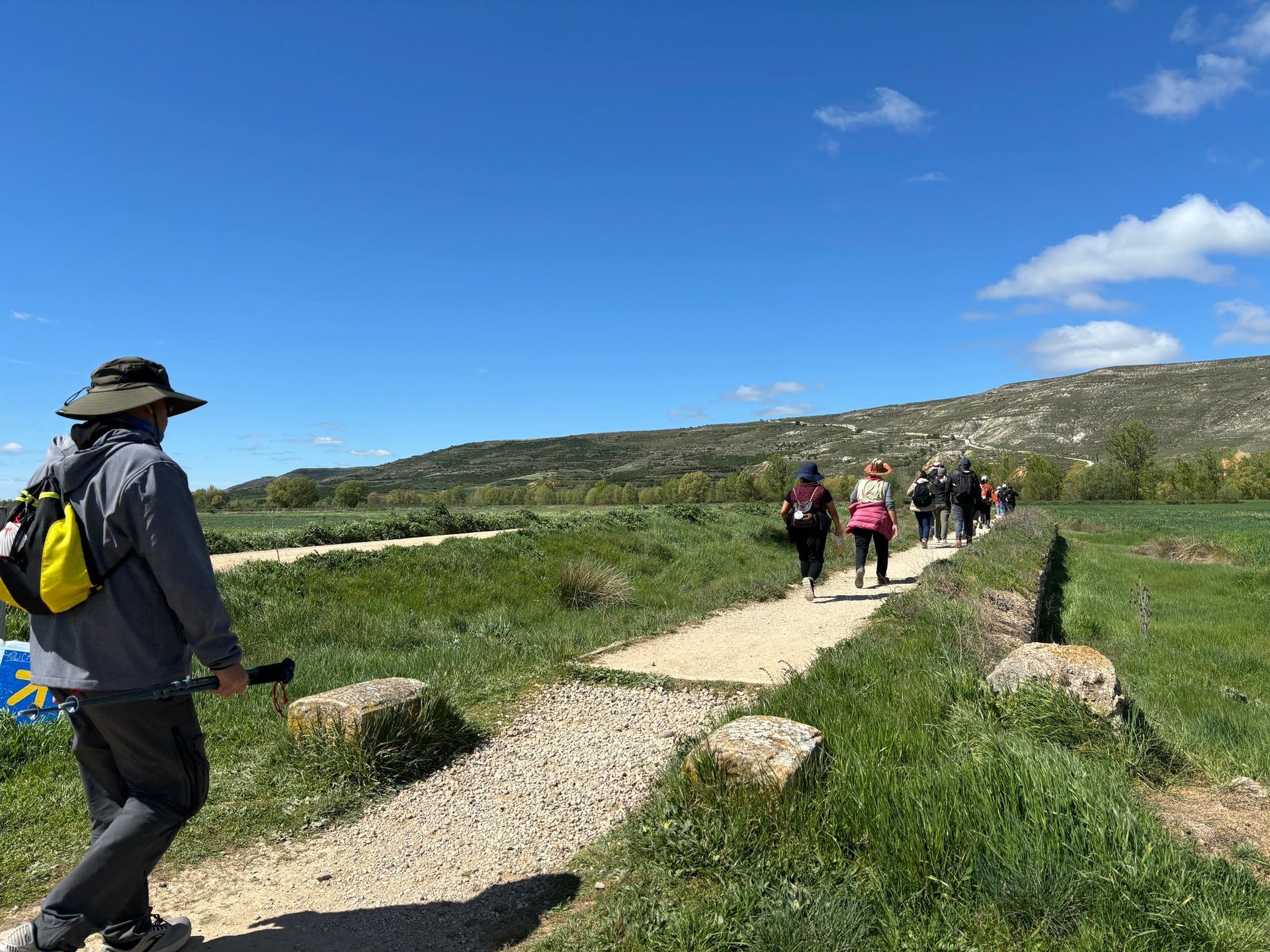 Senderistas caminando por un sendero de grava a través de verdes colinas bajo un cielo azul brillante.