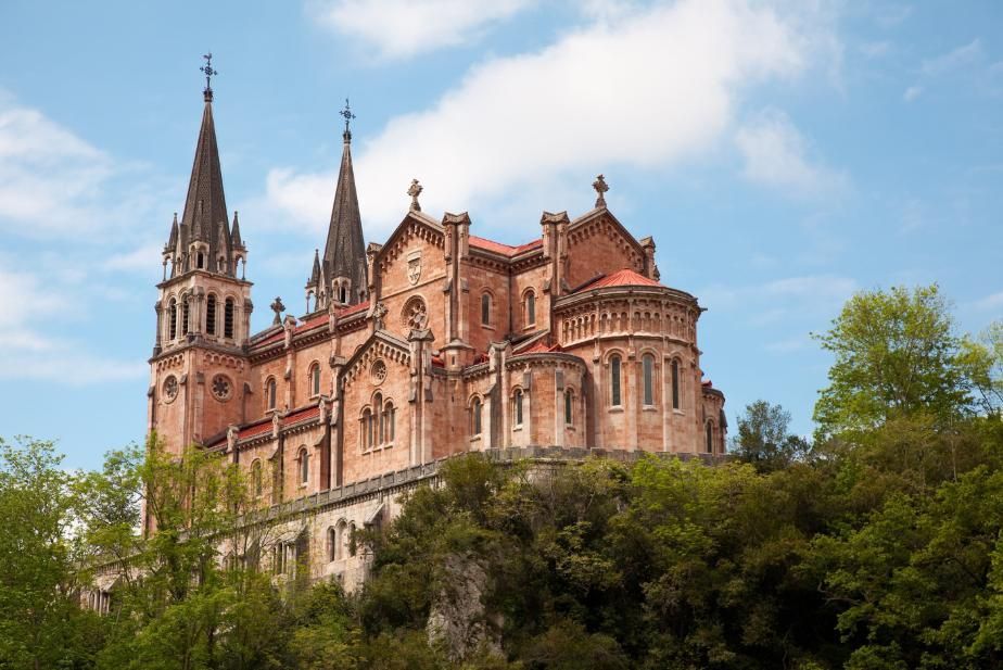 Catedral gótica roja con dos torres en una colina sobre árboles bajo un cielo azul.