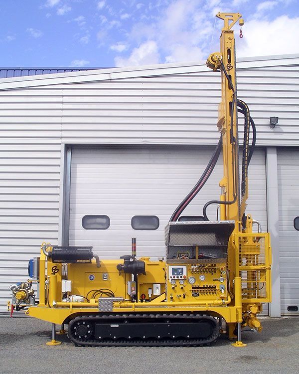 Véhicule de forage jaune garé devant un grand bâtiment blanc en tôle