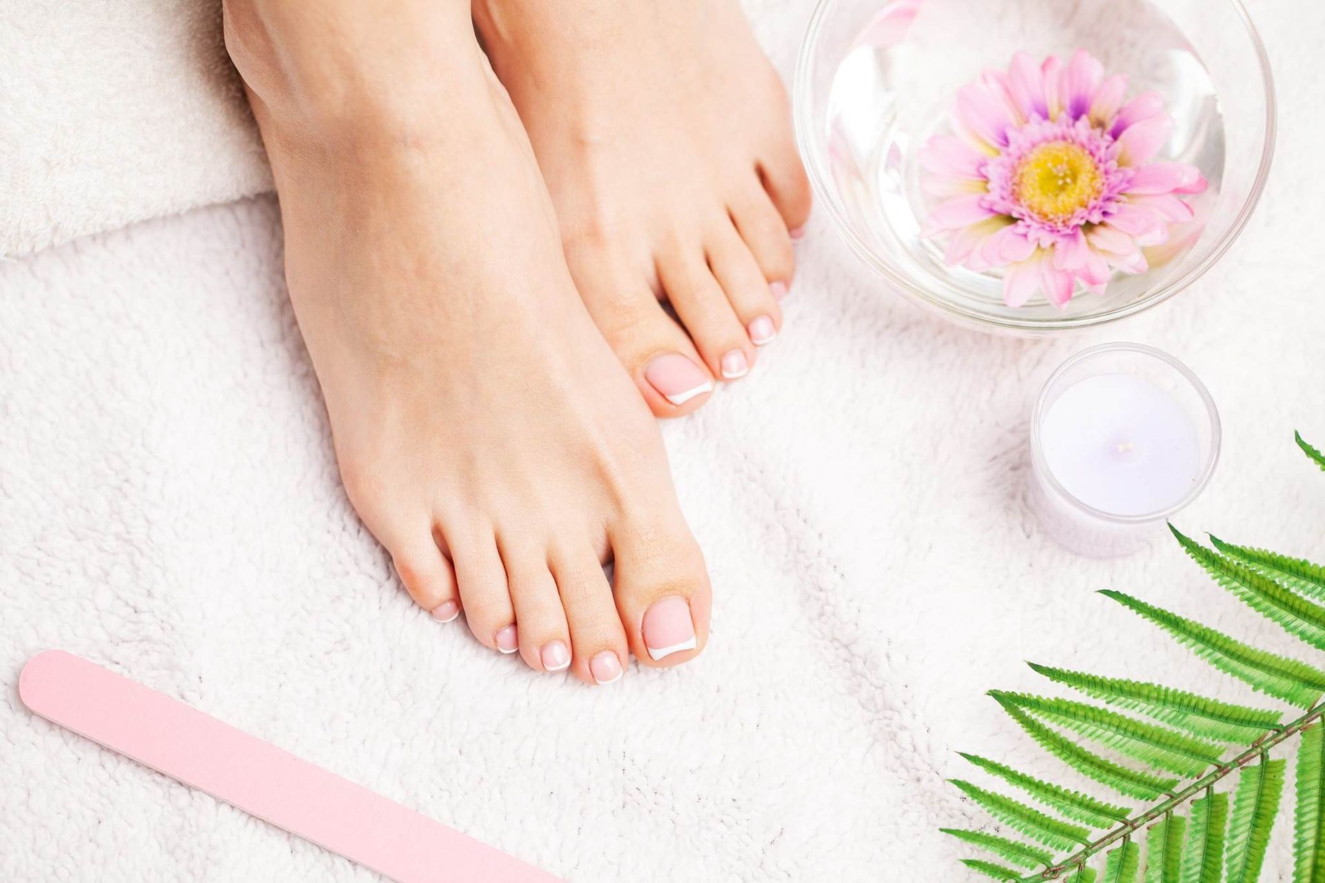 Pies con esmalte de uñas rosa en un ambiente de spa, junto a un jarrón con flores, una vela, una lima de uñas.