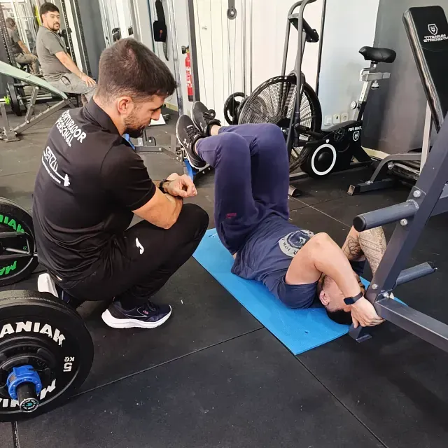 Un hombre en cuclillas junto a un hombre acostado sobre una colchoneta azul en un gimnasio.
